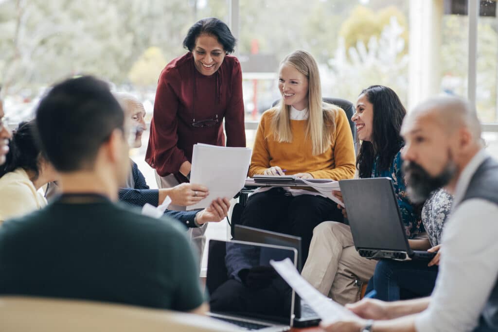 A diverse team looks happy in conversation. They are sitting around with laptops in front of them. Woman of Indian ethnicity leads the team