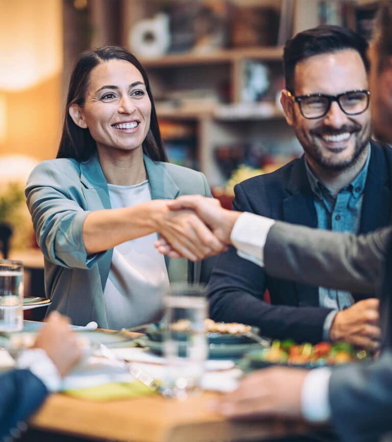 A smiling female in a suit sits beside a male colleague in glasses and she shakes hands with someone during ameeting