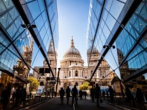 Color image depicting a crowd of people, thrown into silhouette and therefore unrecognisable, walking alongside modern futuristic architecture of glass and steel. In the distance we can see the ancient and iconic dome of St Paul's cathedral, London