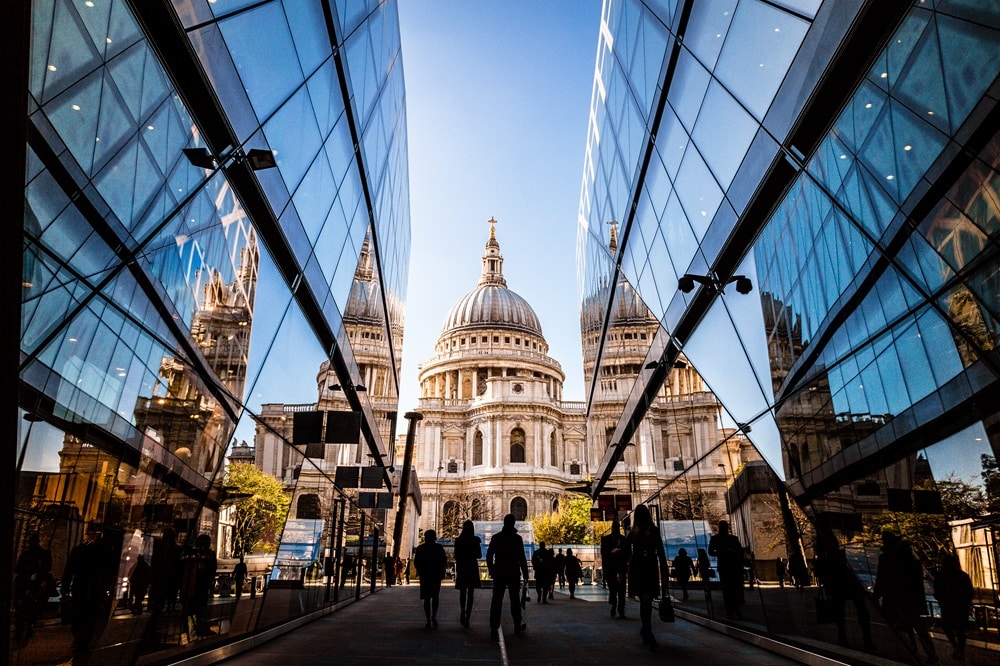 Color image depicting a crowd of people, thrown into silhouette and therefore unrecognisable, walking alongside modern futuristic architecture of glass and steel. In the distance we can see the ancient and iconic dome of St Paul's cathedral, London